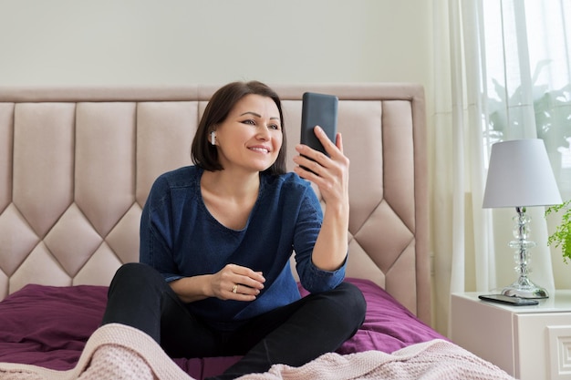 Sonriente mujer de mediana edad con auriculares inalámbricos mirando el teléfono inteligente