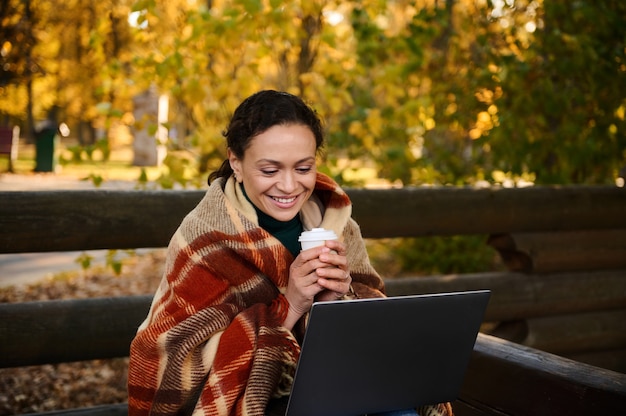 Sonriente mujer de mediana edad envuelta en una cálida manta a cuadros sostiene una taza de papel de café, estudiando, trabajando de forma remota, planificando proyectos sentado en la terraza de madera al aire libre de un acogedor café lejos del ajetreo de la ciudad