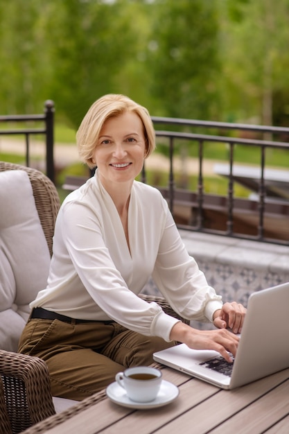 Sonriente mujer de mediana edad rubia agradable teletrabajador sentado en una mesa de madera escribiendo en su computadora portátil