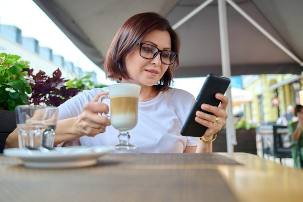 Sonriente mujer de mediana edad sentada en un café al aire libre con una taza de café y mirando el teléfono inteligente