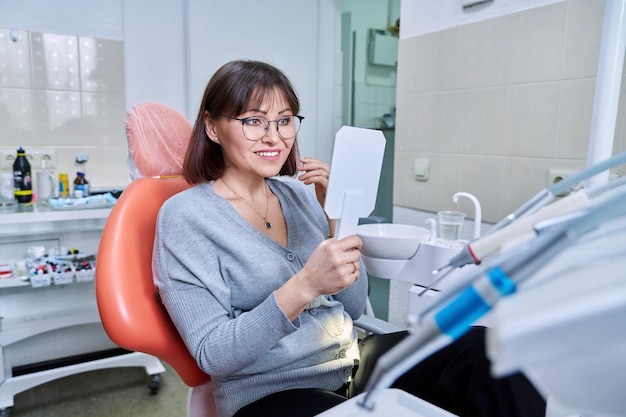 Sonriente mujer de mediana edad en silla dental con espejo mirando sus dientes