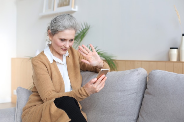 Sonriente mujer de mediana edad sosteniendo el teléfono, usando aplicaciones de dispositivos móviles, mirando la pantalla, mientras está sentado en el sofá.