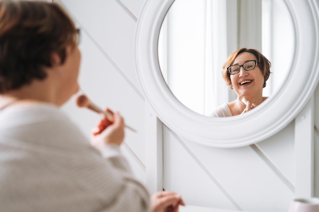 Sonriente mujer de mediana edad y tamaño grande haciendo maquillaje cerca del espejo en casa
