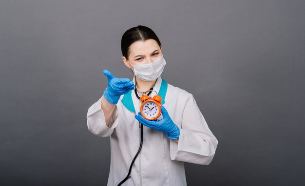 Sonriente mujer médico en máscara apuntando en el reloj, tiempo de vacunación, concepto médico