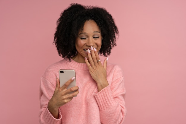 Sonriente mujer milenaria de piel oscura mirando el teléfono móvil en estado de shock