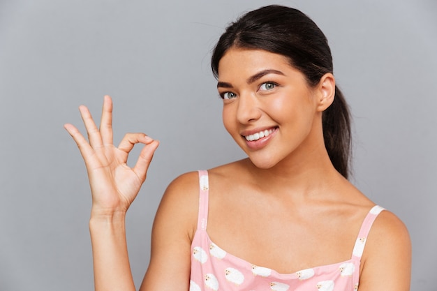 Sonriente mujer morena mostrando signo ok aislado en una pared gris