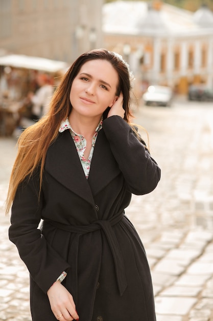 Sonriente mujer morena con pelo largo vistiendo abrigo azul de moda y camisa de algodón posando en día soleado