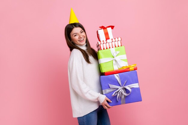 Sonriente mujer morena positiva en cono de fiesta celebrando cumpleaños sosteniendo muchas cajas de regalo