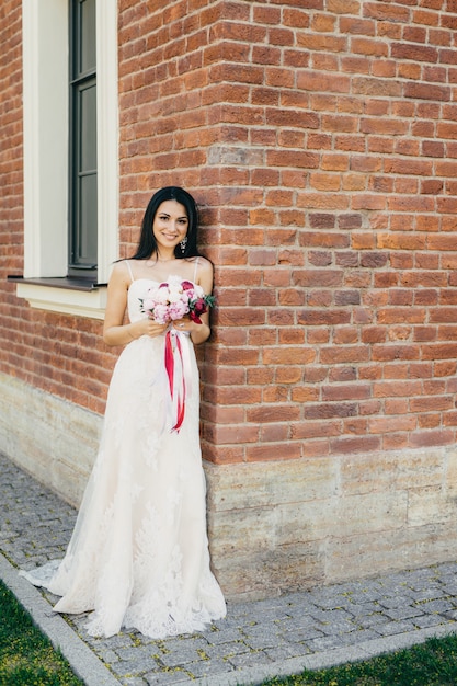 Sonriente mujer morena en vestido de novia blanco largo y hermoso, tiene ramo, se encuentra cerca del edificio de ladrillo