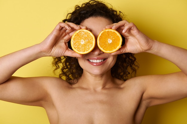 Sonriente mujer musculosa de raza mixta sonrisa saludable y piel facial hidratada brillante, cubriéndose los ojos con dulces mitades de naranja