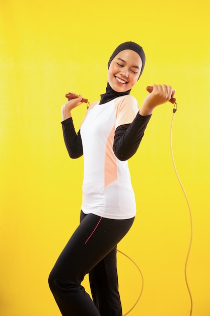 Sonriente mujer musulmana asiática ejerciendo saltar la cuerda sobre un fondo amarillo