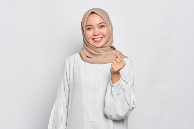 Sonriente mujer musulmana asiática mostrando corazón coreano con dos dedos cruzados aislado sobre fondo blanco.