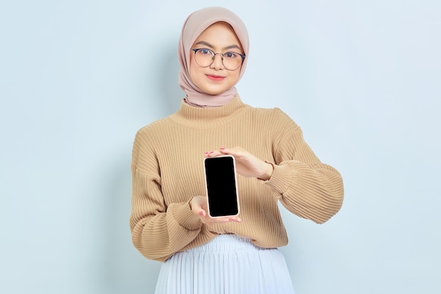 Sonriente mujer musulmana asiática en suéter marrón y hijab mostrando teléfono móvil de pantalla en blanco aislado sobre fondo blanco Concepto de estilo de vida religioso de la gente