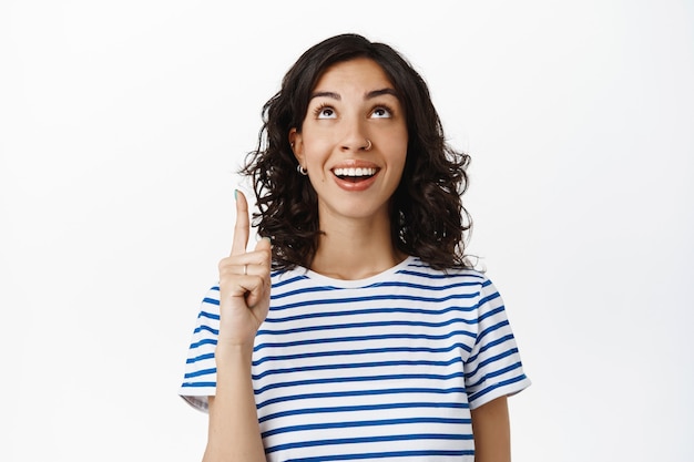 Sonriente mujer natural con cabello oscuro y rizado, nariz perforada, riendo y mirando feliz en el anuncio superior, apuntando con el dedo hacia arriba, mostrando el texto de promoción en blanco