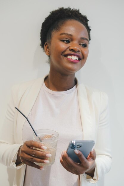 Sonriente mujer de negocios afroamericana con chaqueta blanca bebe café helado con teléfono en cibercafé Trabajo en la industria de negocios y finanzas Mujer independiente disfruta de una bebida relajada