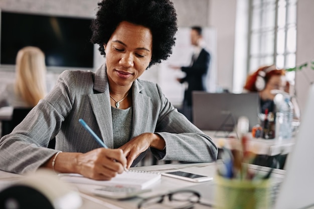 Sonriente mujer de negocios afroamericana trabajando en su escritorio y escribiendo notas en la oficina Hay gente en el fondo