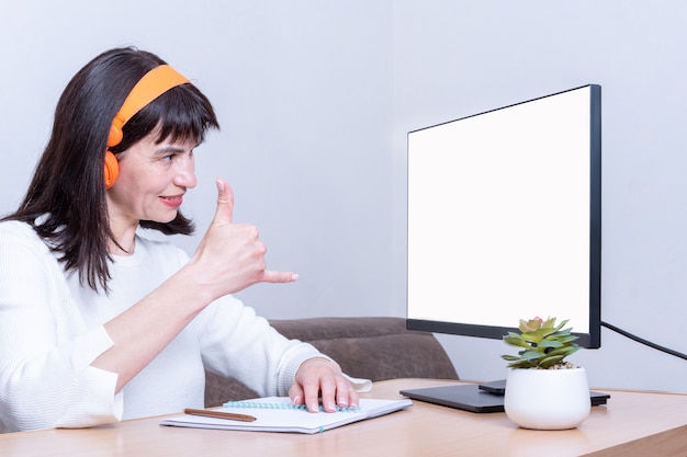 Sonriente mujer de negocios en auriculares mira a un monitor con una pantalla en blanco y muestra un distintivo de llamada, vista lateral, espacio de copia