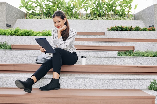 Sonriente mujer de negocios bastante joven sentada en un banco de madera con tablet PC en las manos y sonriendo al frente