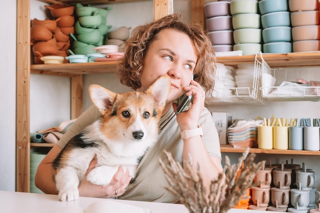 Sonriente mujer de negocios concentrada hablando de teléfono inteligente sosteniendo un lindo corgi de perro tranquilo en el taller Conjunto de platos de silicona para bebés cerca de la pared en los estantes Oficina en casa tomando pedidos en un ambiente confortable