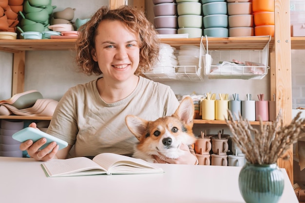 Sonriente mujer de negocios concentrada sosteniendo un teléfono inteligente con un lindo perro corgi en el taller Tienda de platos para bebés de silicona