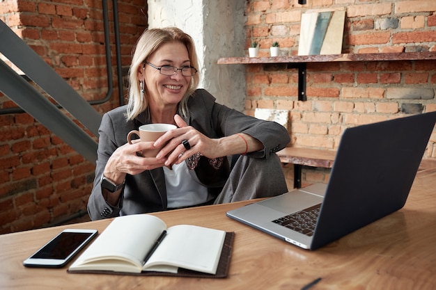 Sonriente mujer de negocios madura tomando café y usando la computadora portátil mientras se comunica con su colega en línea