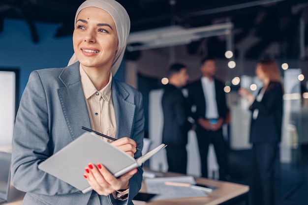 Sonriente mujer de negocios musulmana en hiyab está tomando notas en el libro de trabajo mientras está en el cargo