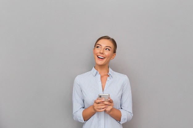 Sonriente mujer de negocios de pie sobre la pared gris de fondo mediante teléfono móvil.