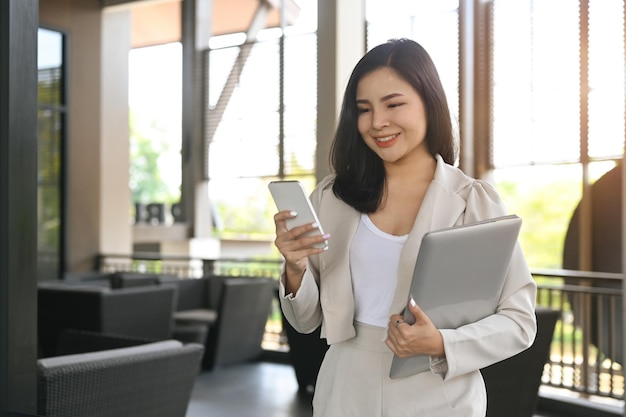 Sonriente mujer de negocios profesional usando teléfono móvil mientras está de pie en la oficina moderna