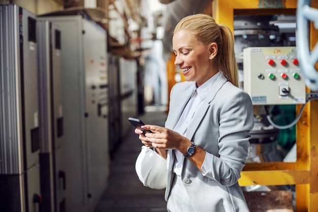 Sonriente mujer de negocios rubia en ropa formal con teléfono inteligente para enviar mensajes de texto mientras está de pie en la planta de energía