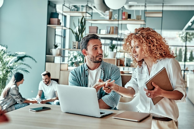 Sonriente mujer de negocios señalando con el dedo en la pantalla del portátil hombre de negocios blanco mirando a sus compañeros de trabajo usando una computadora portátil en el café