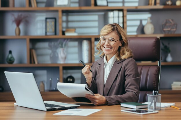 Sonriente mujer de negocios senior con gafas trabajando en la oficina en casa con una computadora portátil y documentos mostrando