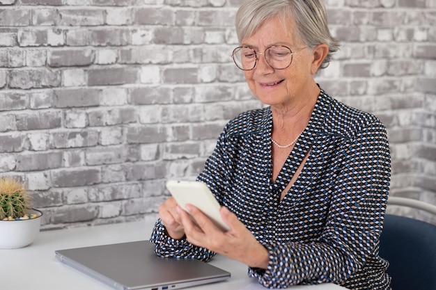 Sonriente mujer de negocios senior segura de sí misma en el escritorio trabajando en una computadora portátil escribiendo un mensaje en un teléfono inteligente