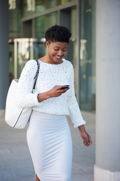 Sonriente mujer negra caminando afuera con teléfono móvil