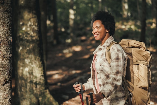 Sonriente mujer negra madura parada sola afuera y cargando una mochila mientras camina por las montañas.