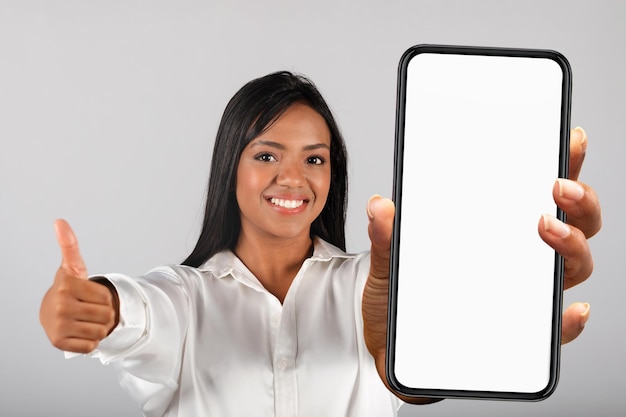 Sonriente mujer negra milenaria confiada en blusa blanca muestra el pulgar hacia arriba y el teléfono inteligente con pantalla vacía