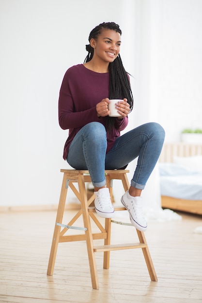 Sonriente mujer negra con taza sentado en heces