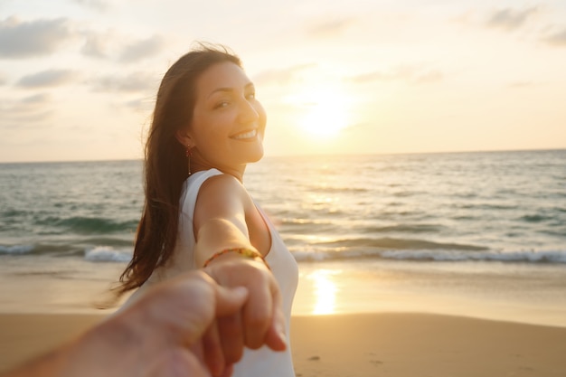 Sonriente mujer de pelo largo corre sosteniendo la mano de su novio a lo largo de la arena de la playa del océano vacío al atardecer