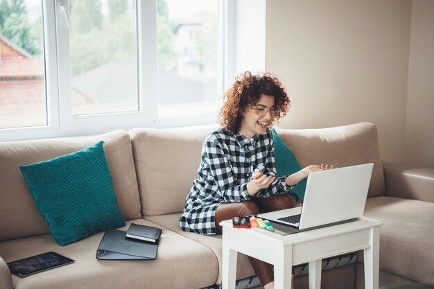 Sonriente mujer de pelo rizado con anteojos sonriendo a la computadora portátil mientras habla con alguien de forma remota