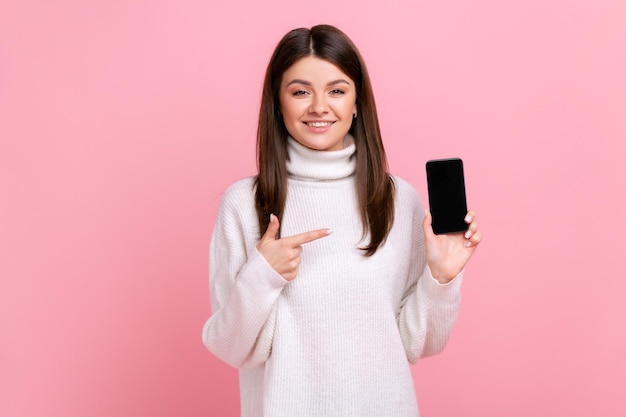 Sonriente mujer positiva apuntando a la pantalla vacía del teléfono inteligente para publicidad o texto promocional