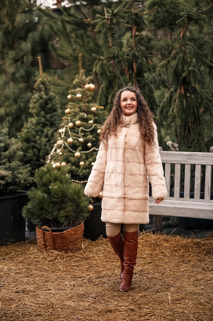 Sonriente mujer rizada feliz en un abrigo de piel está dando vueltas entre los árboles de Navidad en la calle Esperando Navidad y Año Nuevo