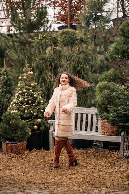 Sonriente mujer rizada feliz en un abrigo de piel está dando vueltas entre los árboles de Navidad en la calle Esperando Navidad y Año Nuevo