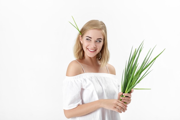 Sonriente mujer rubia con una blusa blanca sosteniendo una cebolla verde sobre un fondo blanco. Salud y vitaminas