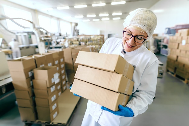 Sonriente mujer rubia empleada en uniforme estéril y con anteojos poniendo cajas en la pila. Interior de la fábrica de alimentos.