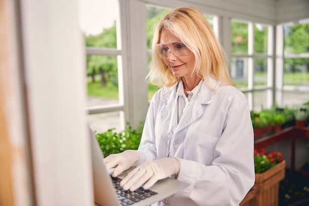 Sonriente mujer rubia madura enfocada en guantes de látex y gafas de seguridad escribiendo en su computadora portátil