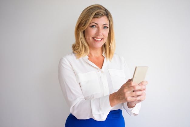 sonriente mujer rubia de mediana edad atractiva mirando a cámara y con smartphone