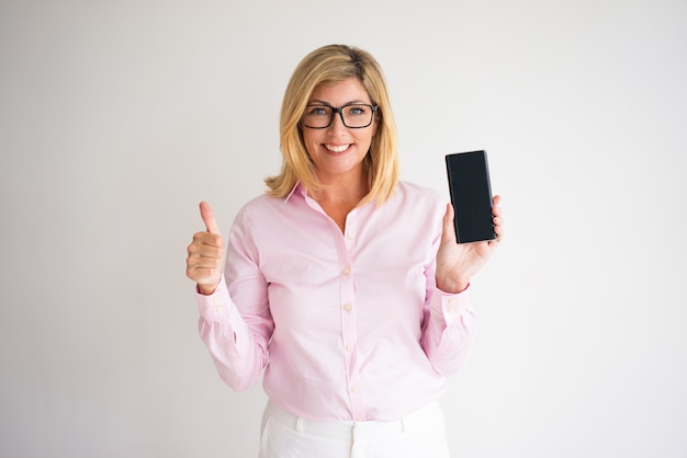 sonriente mujer rubia de mediana edad atractiva mostrando el pulgar hacia arriba y el teléfono inteligente