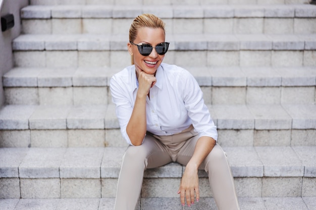 Sonriente mujer rubia de moda atractiva glamorosa con gafas de sol con la mano en el mentón sentado al aire libre en las escaleras.