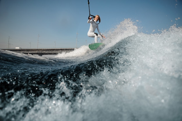 Sonriente mujer rubia en traje de baño blanco de pie en el wakeboard sosteniendo una cuerda en el puente y el cielo