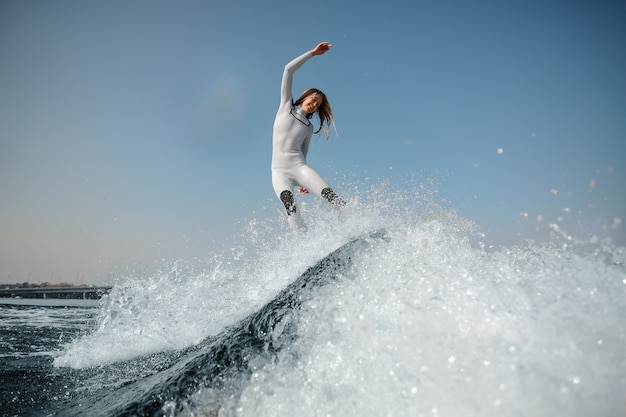 Sonriente mujer rubia en traje de baño blanco saltando sobre el wakeboard verde sobre las rodillas dobladas