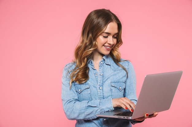 Foto sonriente mujer rubia vestida con camisa de mezclilla con ordenador portátil sobre pared rosa